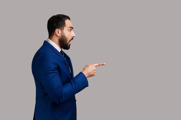 Vue latérale d'un homme barbu regardant la caméra avec une expression étonnée surprise, pointant vers l'espace de copie, portant un costume de style officiel. Tourné en studio intérieur isolé sur fond gris.