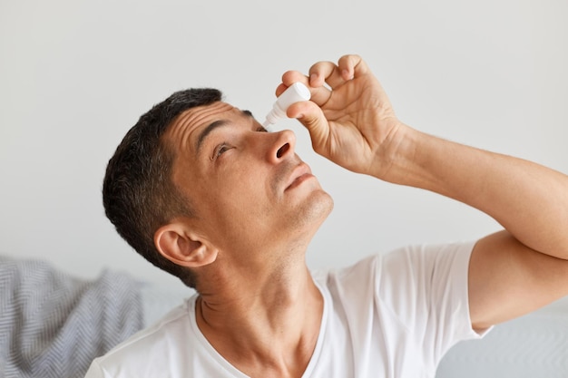 Vue latérale d'un homme brune caucasienne malade portant un t-shirt blanc, posant une pièce lumineuse alors qu'il était assis sur un canapé gris, ayant des problèmes avec les yeux, utilisant des gouttes pour les yeux pour traiter.