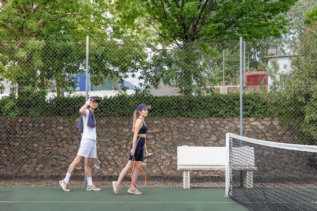 Vue latérale d'un homme et d'une femme vêtus de sports arrivant sur un court de tennis pour jouer un match avec des raquettes à portée de main