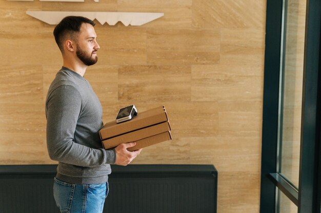 Vue latérale d'un homme de messagerie tenant des boîtes de pizza et un terminal sans fil POS de paiement sans contact debout dans le hall d'entrée d'un appartement ou d'un immeuble de bureaux