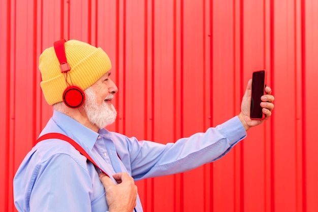 Vue latérale d'un homme senior heureux prenant un selfie tout en écoutant de la musique avec un casque sur fond rouge Hipster vieil homme s'amusant