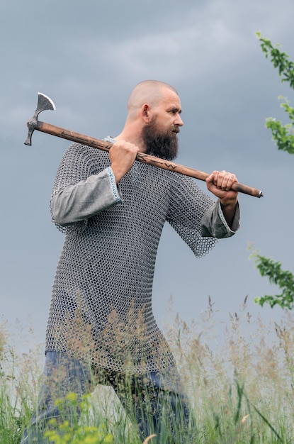 Photo vue latérale d'un homme en vêtements vikings médiévaux, casque en métal et chaînes de fer menaçant avec une hache.