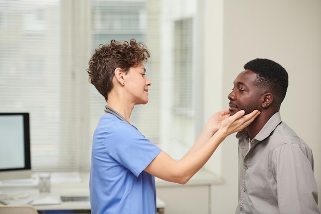 Vue latérale horizontale d'une médecin portant un uniforme bleu vérifiant la taille des glandes lymphatiques des patients afro-américains