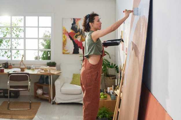 Vue latérale d'un jeune artiste en vêtements décontractés tenant un plateau avec de la peinture et un pinceau