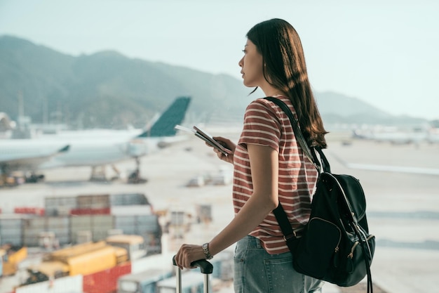 vue latérale d'une jeune étudiante de voyage explore le monde pendant les vacances de printemps en attendant le vol dans la porte d'embarquement debout près des avions de fenêtre sur la piste à l'arrière. dame tenir un téléphone portable à l'aéroport de hong kong