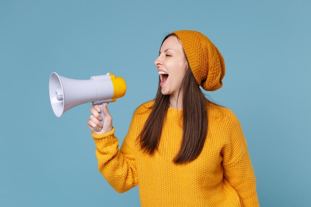 Vue latérale d'une jeune femme brune fille en pull jaune et chapeau posant isolé sur fond bleu portrait en studio. Concept de mode de vie des émotions sincères des gens. Maquette de l'espace de copie. Crier dans le mégaphone.