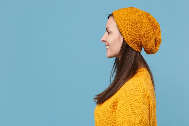 Vue latérale d'une jeune femme brune souriante en chapeau pull jaune posant isolé sur fond bleu portrait en studio. Concept de mode de vie des émotions sincères des gens. Maquette de l'espace de copie. Regarder de côté.