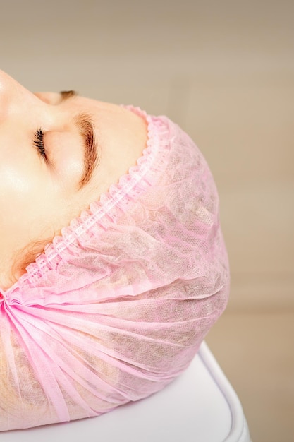 Vue latérale d'une jeune femme caucasienne dans une casquette médicale allongée les yeux fermés en attente de la procédure cosmétique dans un salon de beauté.