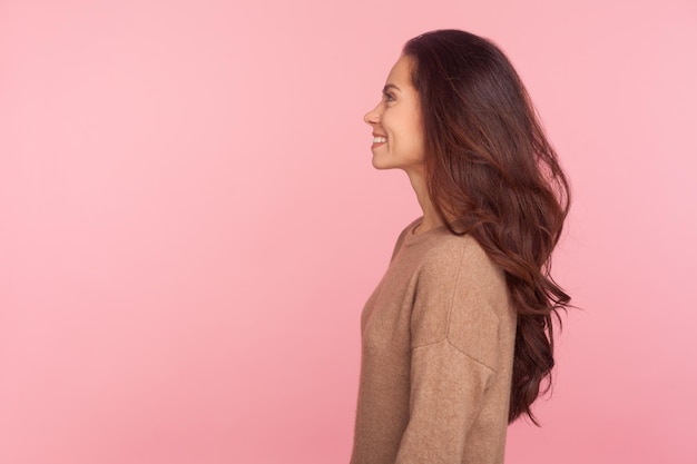 Vue latérale d'une jeune femme heureuse avec de longs cheveux ondulés brune souriant joyeusement, exprimant l'optimisme, les émotions positives, la beauté féminine et le concept de mode. studio d'intérieur tourné isolé sur fond rose