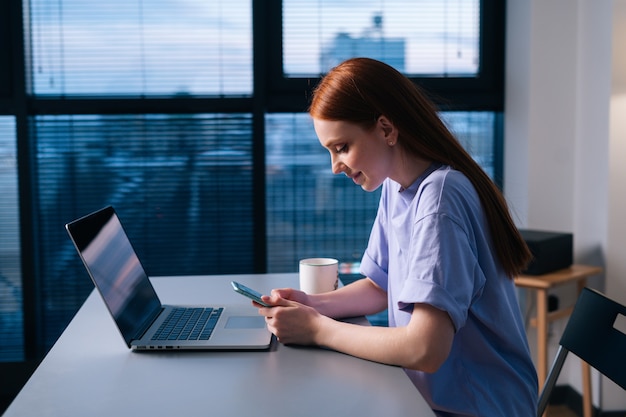 Vue latérale d'une jeune femme joyeuse utilisant un téléphone portable assis au bureau avec un ordinateur portable près de la fenêtre