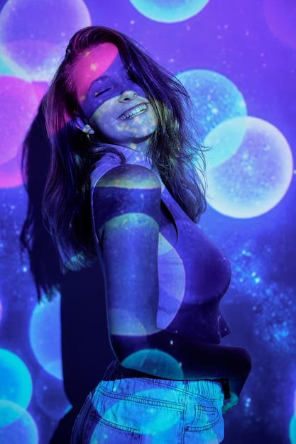Vue latérale d'une jeune femme optimiste aux cheveux longs souriant joyeusement avec les yeux fermés et touchant le ventre tout en dansant sous la projection d'éclairs colorés