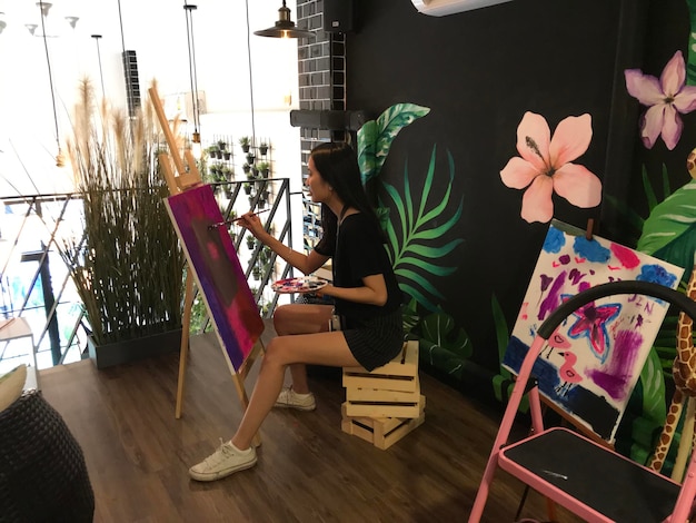 Photo vue latérale d'une jeune femme peignant sur toile alors qu'elle est assise sur des caisses à la maison