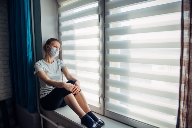Vue latérale jeune femme en portant un masque de protection assis sur le rebord de la fenêtre à la maison, regardant à l'extérieur. Auto-isolement, prévention de la pandémie de coronavirus en quarantaine. Concept d'épidémie et de prévention