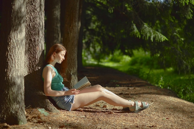 Vue latérale d'une jeune femme rousse blanche en haut vert et short bleu lisant dans la forêt