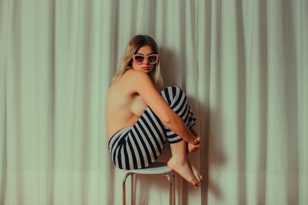 Vue latérale d'une jeune femme sans chemise portant des lunettes de soleil assise sur une chaise par un rideau