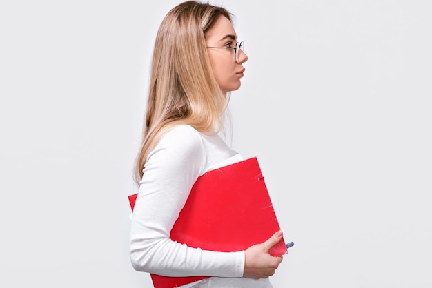 Vue latérale d'une jeune femme sérieuse portant un chemisier blanc et des lunettes rondes à la mode avec un dossier rouge à la main regarde loin vers l'espace de copie vierge Employée de bureau européenne posant sur un mur de studio blanc