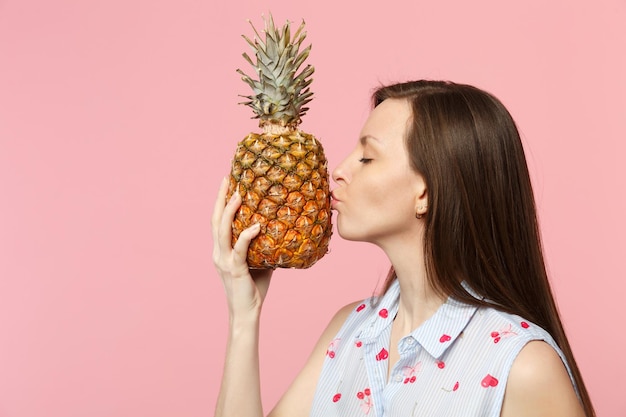 Vue latérale d'une jeune femme en vêtements d'été tenant embrassant des fruits d'ananas mûrs frais isolés sur fond de mur pastel rose en studio. Les gens vivent un style de vie vivant et se détendent. Maquette de l'espace de copie.