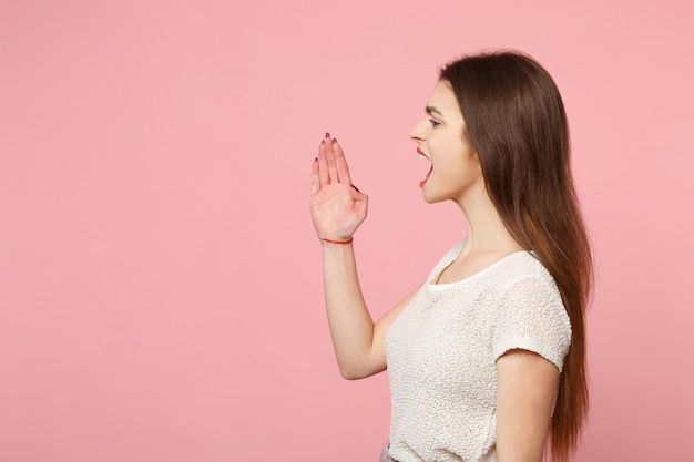 Vue latérale d'une jeune femme en vêtements légers décontractés posant isolé sur fond de mur rose pastel, portrait en studio. Concept de style de vie des gens. Maquette de l'espace de copie. Crier, avec un geste de la main près de la bouche.