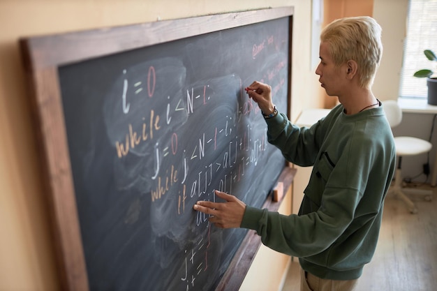 Vue latérale d'un jeune homme blond en tenue décontractée écrivant des formules ou des équations sur un tableau noir avec de la craie tout en expliquant quelque chose en leçon