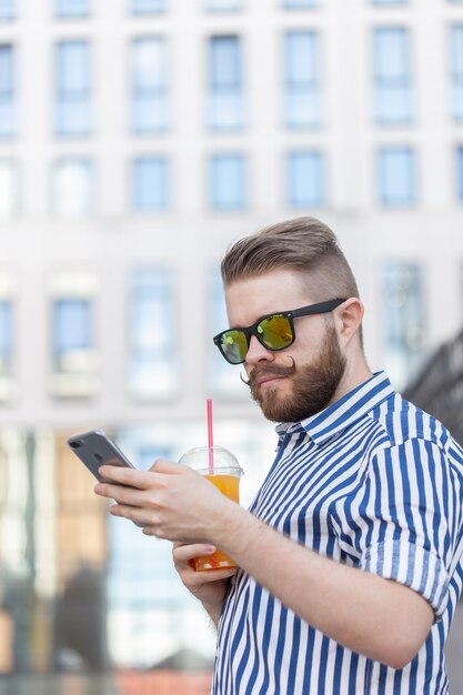 Vue latérale d'un jeune homme élégant hipster avec des lunettes et une moustache et du jus dans ses mains en discutant dans les réseaux sociaux à l'aide d'un smartphone et Internet sans fil par une chaude journée d'été.