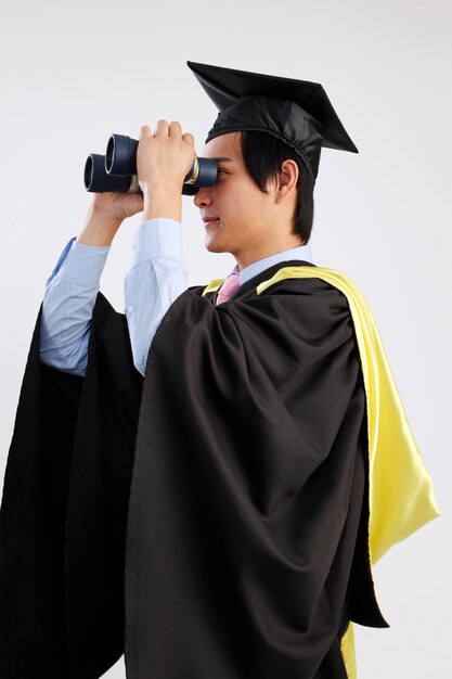 Photo vue latérale d'un jeune homme souriant en robe de graduation utilisant des jumelles sur fond blanc