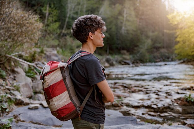 Vue latérale sur le jeune voyageur caucasien avec sac à dos debout dans un endroit rural à la rivière