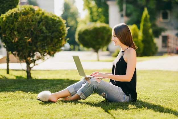 Vue latérale d'une jolie fille souriante dans des vêtements à la mode, assis sur l'herbe et à l'aide d'un ordinateur portable. Dans le parc.