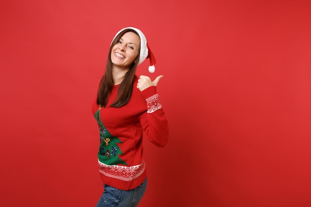 Vue latérale d'une joyeuse jeune fille de Santa en pull tricoté, chapeau de Noël pointant le pouce de côté isolé sur fond de mur rouge. Bonne année 2019 concept de fête de vacances célébration. Maquette de l'espace de copie.