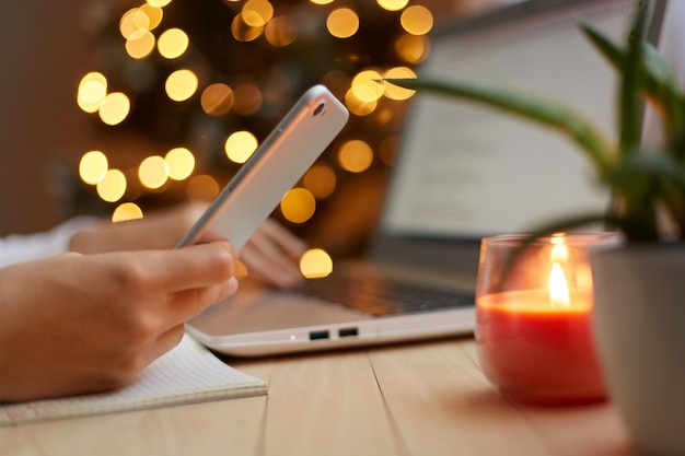 Vue latérale de la main d'une femme tenant un téléphone portable dans les mains d'une femme inconnue utilisant un téléphone portable naviguant sur Internet avec un sapin de Noël flou sur fond