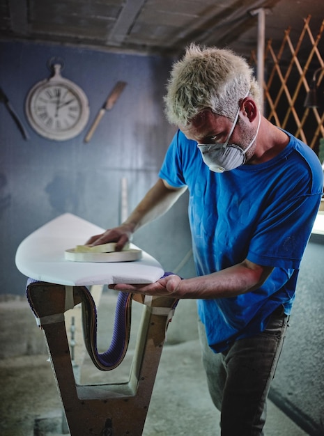 Vue latérale d'un maître à cheveux blonds concentrés dans un masque respiratoire polissant une planche de surf blanche placée sur des supports de façonnement dans un atelier