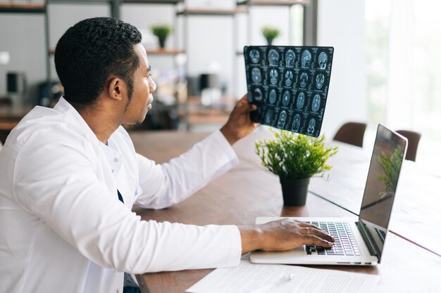 Vue latérale d'un médecin noir afro-américain en manteau blanc analysant l'histoire de la maladie du patient à l'aide d'un scanner de tête cérébrale IRM, travaillant sur un ordinateur portable. Concept de médecine et de soins de santé.