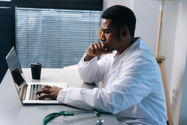 Vue latérale d'un médecin de sexe masculin noir confus portant un uniforme de blouse blanche pensant en regardant un écran d'ordinateur portable assis au bureau