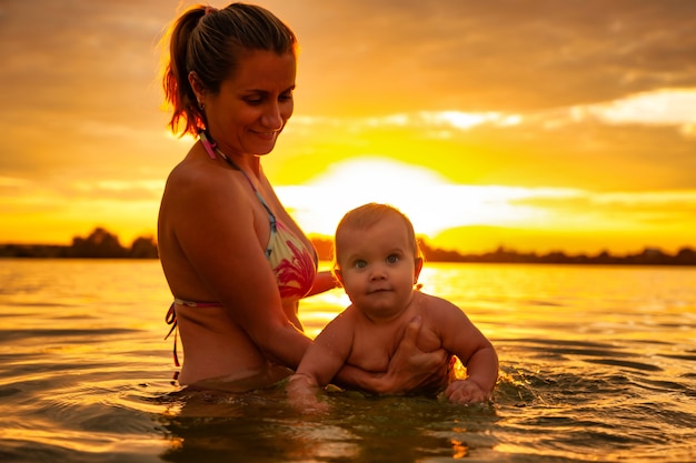 Vue latérale d'une mère caucasienne heureuse nageant avec un petit bébé souriant mignon en mer. Belle famille profitant de l'été à l'extérieur pendant un fantastique coucher de soleil orange vif. Notion de famille.