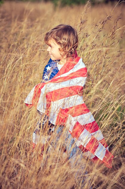 Vue latérale d'un mignon garçon préadolescent enveloppé dans le drapeau national des États-Unis d'Amérique debout dans une prairie de seigle