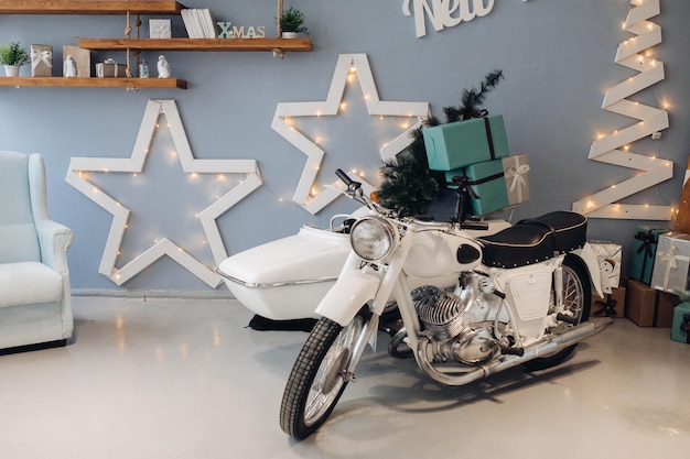 Vue latérale d'une moto rétro blanche chargée de cadeaux de Noël verts emballés du père Noël et du sapin de Noël. Moto avec des cadeaux dans une chambre décorée.