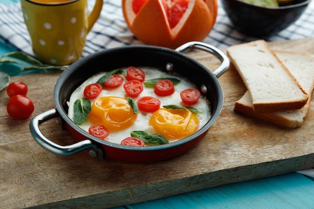 Vue latérale des œufs au plat avec des tomates dans une poêle décorée de feuilles avec des tranches de pain sur une planche à découper avec une tasse de jus d'orange pamplemousse sur un tissu à carreaux sur fond bleu