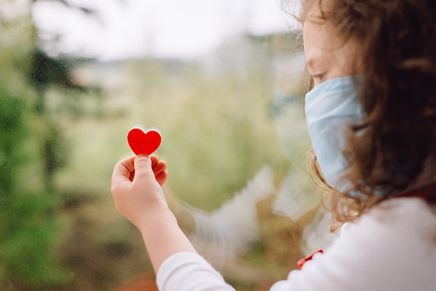 Vue latérale d'une petite fille d'âge préscolaire en portant un masque protecteur, assis sur le seuil à la maison, tenant un petit coeur rouge. Restez à la maison pour la prévention de la pandémie de coronavirus en quarantaine. Quarantaine à domicile