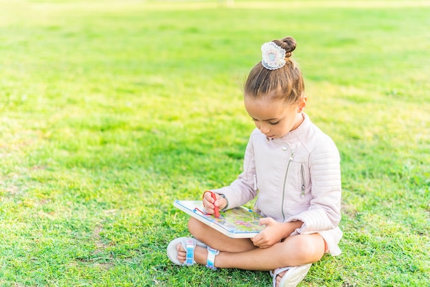 Vue Latérale D'une Petite Fille Assise Sur L'herbe Dans Un Parc Jouant Avec Une Tablette Graphique.