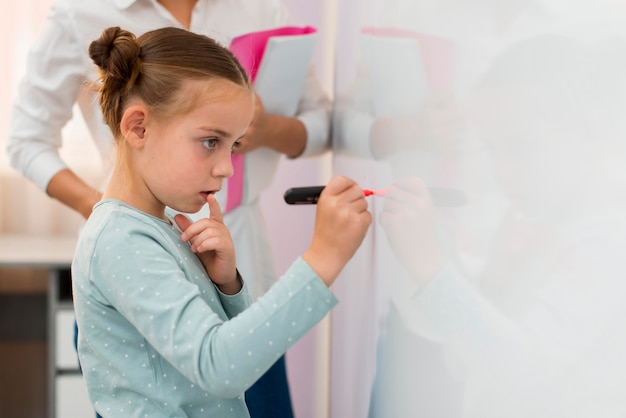 Vue latérale petite fille écrivant sur un tableau blanc à côté de son professeur