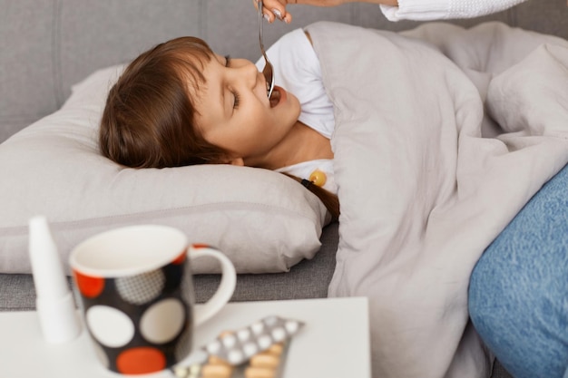 Vue latérale d'une petite fille malsaine allongée sur la toux, d'une mère ou d'un médecin prenant soin de son enfant malade, donnant des médicaments, traitant la grippe, buvant du sirop en toussant.