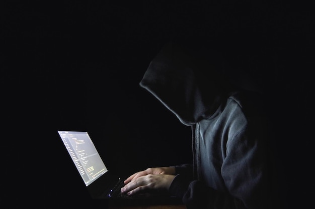 Vue latérale d'un pirate informatique portant une chemise à capuche tout en utilisant un ordinateur portable sur un fond noir