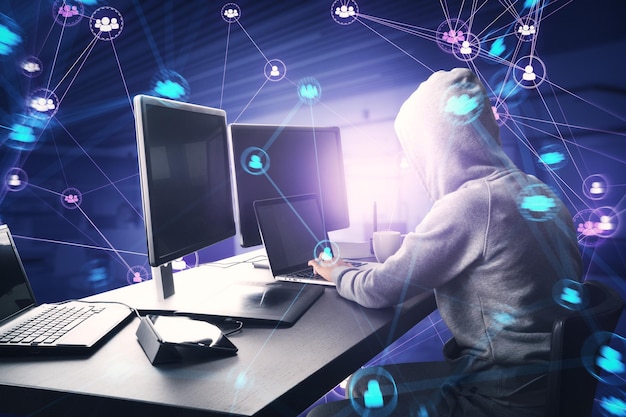 Vue latérale d'un pirate informatique en sweat à capuche utilisant des ordinateurs au bureau avec des icônes d'équipe de personnes numériques connectées sur fond intérieur de bureau violet foncé et flou Communauté de réseau numérique et concept de médias sociaux