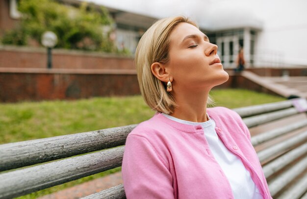 Vue latérale portrait d'une belle jeune femme blonde se relaxant à l'extérieur sur le banc avec les yeux fermés portant un t-shirt blanc et une veste rose dans la rue de la ville Jolie femme se reposant dehors au printemps