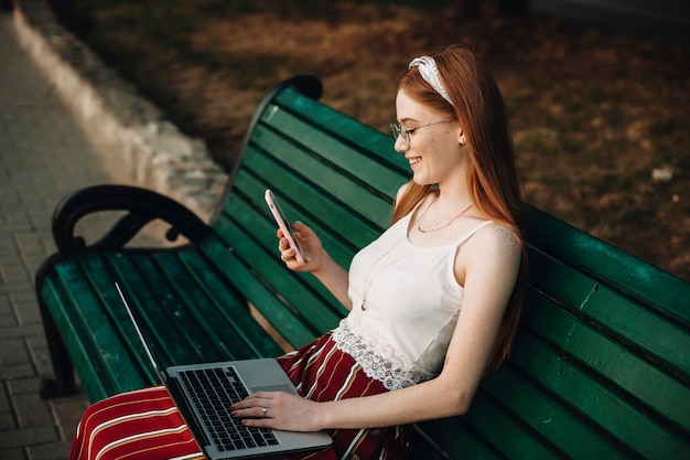 Vue latérale portrait d'une belle jeune femme regardant un smartphone avec un ordinateur portable sur ses jambes. Influenceuse aux cheveux roux et taches de rousseur assis sur un banc.