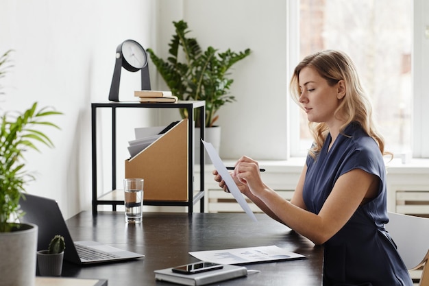 Vue latérale portrait d'une femme d'affaires blonde signant un document tout en travaillant au bureau dans un bureau à domicile.