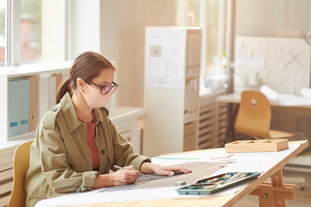 Vue latérale portrait de femme architecte portant un masque alors qu'il était assis au bureau de dessin au soleil,