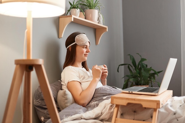 Vue latérale portrait d'une femme calme aux cheveux bruns en t-shirt blanc et masque de sommeil tenant une tasse de café ou de thé dans les mains en regardant l'écran de l'ordinateur portable en regardant un film