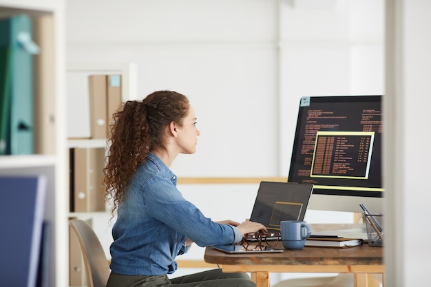 Vue latérale portrait de femme développeur informatique à l'aide de l'ordinateur lors du codage dans l'intérieur de bureau blanc moderne, espace copie