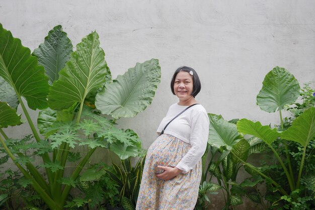 Vue latérale portrait d'une femme enceinte asiatique heureuse sur fond de nature verte, regardant la caméra, concept COVID-19