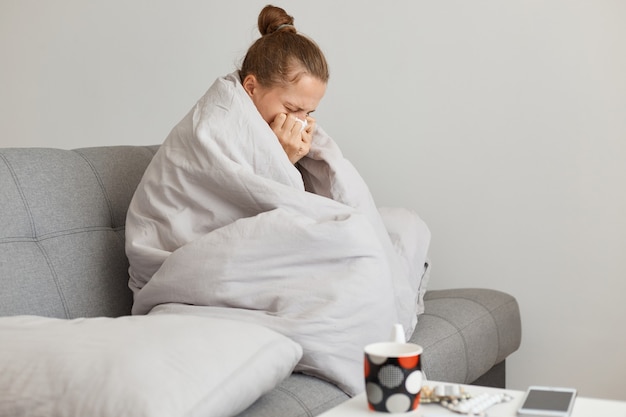 Vue latérale portrait d'une femme malade avec un chignon assise sur un canapé enveloppé dans une couverture, souffrant d'écoulement nasal et de température élevée, attrapant froid, posant dans la chambre à la maison.